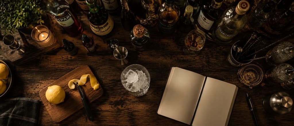 An overhead view of the bar at the moment of decision — a glass with ice, citrus, and an open notebook waiting to be filled.